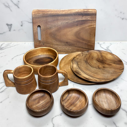 A set of handcrafted wooden tableware, including a rectangular cutting board with a handle, two stacked wooden bowls, two stacked wooden mugs with handles, two round serving plates, and four small round bowls, all displayed on a white marble surface.